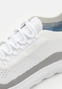 Zapatilla blanca y gris con parte superior de punto transpirable, cierre con cordones y suela texturizada. Presenta la marca GEOX en el lateral.