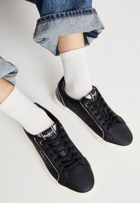 Sneaker sintetiche nere con punta rotonda, accenti beige a contrasto sui lati e lacci classici. Abbinate a calze bianche a coste.
