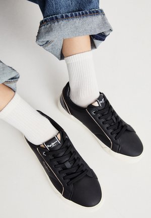 Černé syntetické tenisky s kulatou špičkou, kontrastními béžovými akcenty po stranách a klasickým šněrováním. Spárovány s bílými žebrovanými ponožkami.