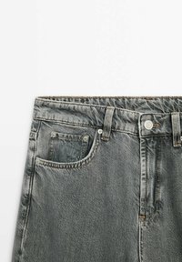 Jean en denim gris avec un design classique à cinq poches, fermeture à bouton et accents de couture subtils. Le matériau semble texturé et usé.