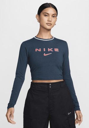 Top corto de manga larga de color navy con cuello de rayas blancas y rojas. Presenta un audaz logo rojo de "NIKE" y su característica marca. Combinado con pantalones negros de cintura alta.