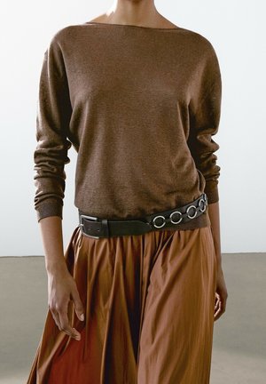 Person iført en brun langærmet sweater, der er stoppet ned i et brunt nederdel med et sort bælte med store metalkringler.