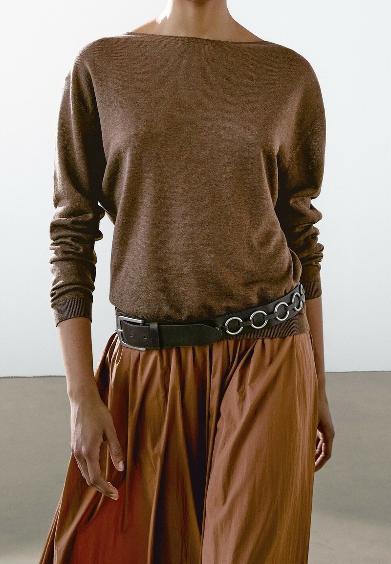 Personne portant un pull marron à manches longues rentré dans une jupe marron avec une ceinture noire ornée de grands anneaux métalliques.