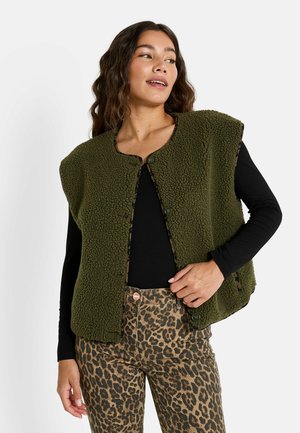 Gilet en polaire vert olive avec un col rond, des boutons sur le devant, et une texture douce, porté sur une chemise noire à manches longues et un pantalon à imprimé léopard.