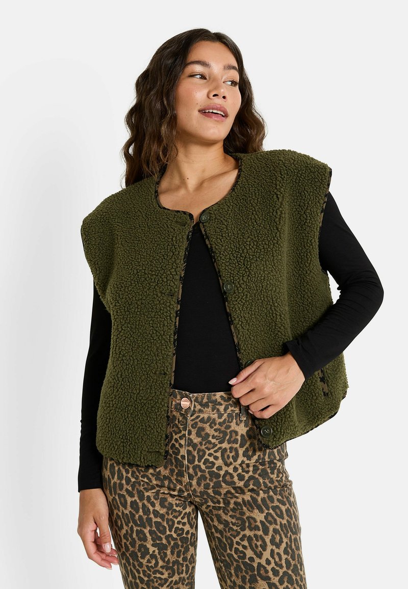 Gilet en polaire vert olive avec un col rond, des boutons sur le devant, et une texture douce, porté sur une chemise noire à manches longues et un pantalon à imprimé léopard.