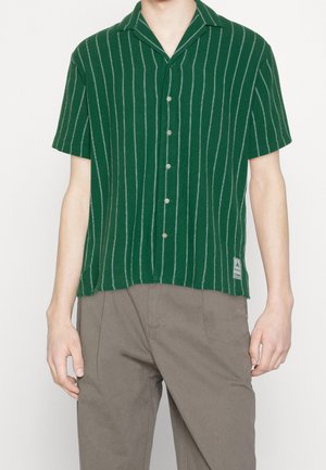 Camicia verde a maniche corte con righe verticali bianche, chiusura frontale con bottoni e colletto a punta. Presenta un tessuto texturizzato e una patch con logo all'orlo.