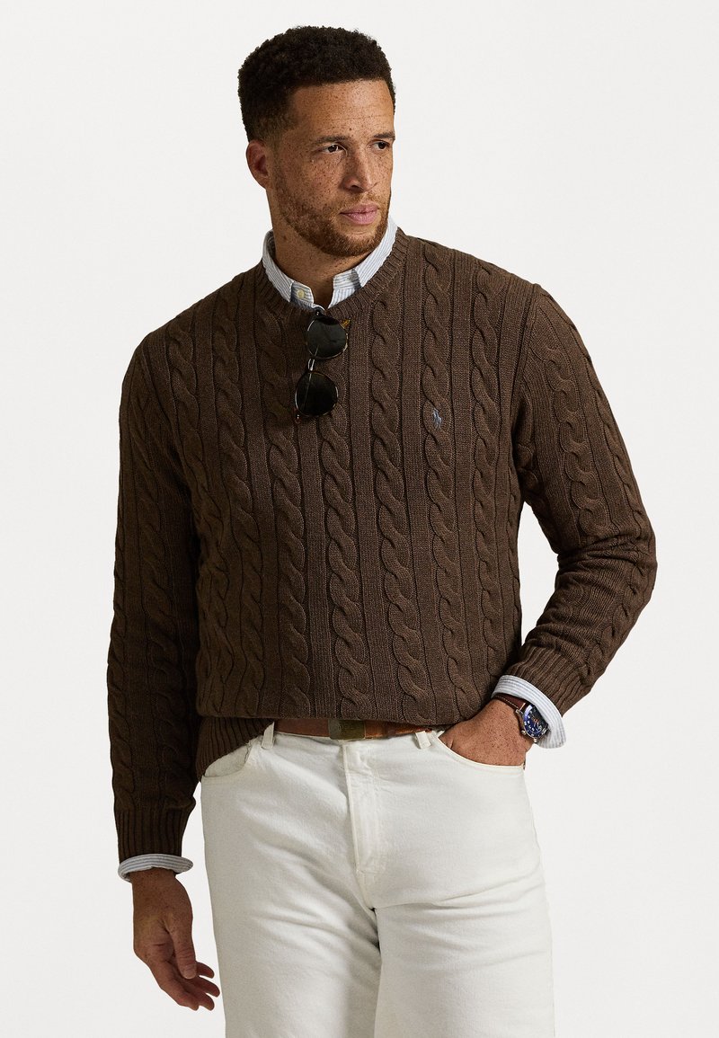 Bruin kabelgebreid sweater met ronde hals, gedragen over een lichtblauw shirt met kraag. Gekozen met lichtgekleurde broek en zonnebril.