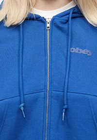 Κοντινό πλάνο ατόμου που φοράει μπλε φούτερ με φερμουάρ και κορδόνια, με μερικώς ορατό ανοιχτόχρωμο κείμενο στην αριστερή πλευρά του στήθους.