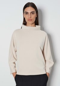 Crème-kleurige top met een hoge, opgevouwen kraag, losse pasvorm, verlaagde schouders en lange mouwen, gemaakt van een gladde, zachte stof.