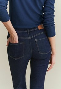Jean en denim bleu foncé au design ajusté, avec deux poches arrière et des coutures dorées contrastantes. Texture lisse avec une étiquette en cuir au niveau de la taille.