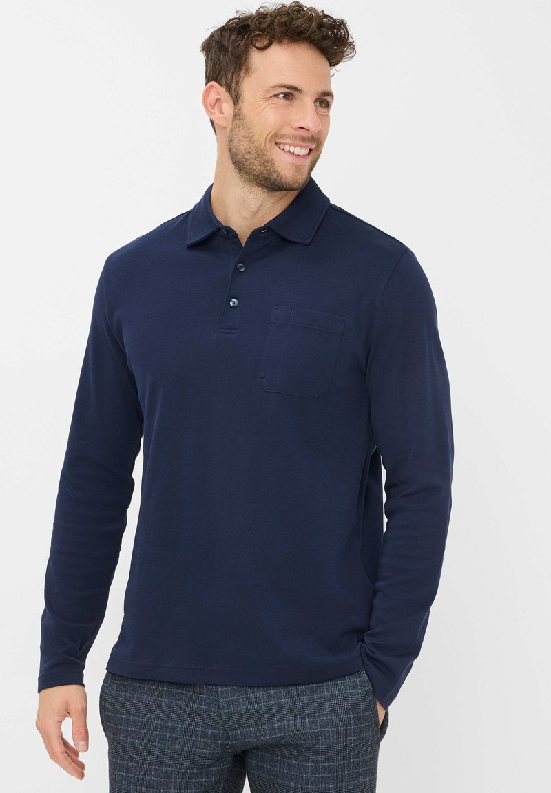 Mann lächelt, trägt ein navyblaues Langarm-Polohemd mit Fronttasche und graue karierte Hose, steht vor einem weißen Hintergrund.