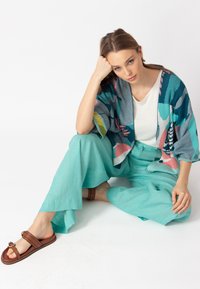 Atsipalaidavęs kimono švarkas mėlynos spalvos su gėlių raštu, derinamas su šviesiai mėtinėmis plačiomis kelnėmis ir rudos spalvos šlepetėmis su dviem dirželiais.