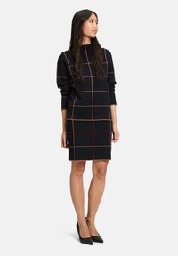Schwarzes Kleid mit Karomuster in Orange. Hat lange Ärmel und einen hohen Ausschnitt. Glatte Textur und taillierte Form, endet oberhalb des Knies.