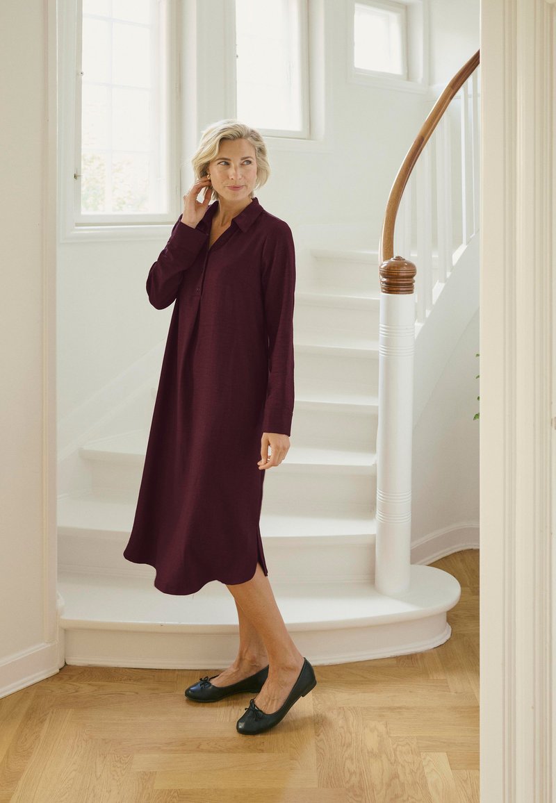 Burgunderrotes Langarmkleid mit Kragen und Seitenschlitzen, kombiniert mit schwarzen Ballerinas; im Hintergrund eine hölzerne Wendeltreppe.