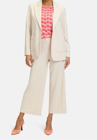 Beige skräddarsydd blazer med två frontfickor över en rosa mönstrad topp, tillsammans med ljusbeiga vida byxor och persikofärgade klackar.