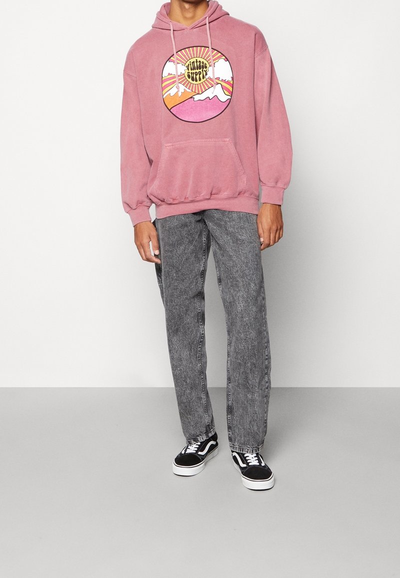 Personne portant un sweat à capuche rose avec un graphisme de montagne et du texte, un jean gris délavé à l'acide et des baskets noires et blanches.