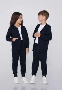 Dwoje dzieci nosi pasujące granatowe bluzy z kapturem na zamek i dresy, wykonane z miękkiego materiału, w połączeniu z białymi koszulkami i trampkami.