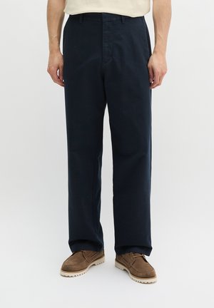 Mężczyzna ubrany w ciemnogranatowe spodnie o prostych nogawkach i brązowe sznurowane buty, stojący na tle jednolitego jasnego tła.