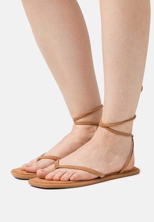 Sandales plates en cuir marron avec de fines lanières autour de la cheville et un design minimaliste. La semelle est plate avec une texture lisse, mettant en valeur des pieds nus.