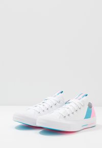 Zapatillas de lona blancas con detalles en azul y rosa, suela de goma, puntera redonda y ojales de metal. Diseño bajo con paneles de color contrastante.