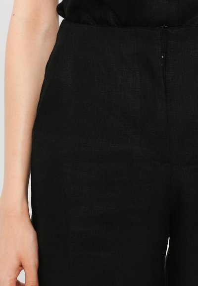 Black linen pants with a fitted waist and textured surface, featuring a side zipper and flat front design, shown with a hand on the hip.