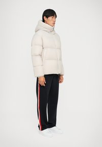Puffjacka i ljusbeige med hög krage, med vadderade sektioner och dragkedja. Kombineras med marinblå byxor och vita sneakers.