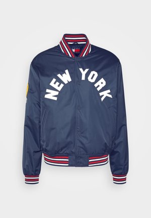 Person trägt eine dunkelblaue New York Varsity-Jacke, ein schwarzes T-Shirt, hellblaue Jeans mit Muster, weiße Socken und dunkelblaue Schlappen.