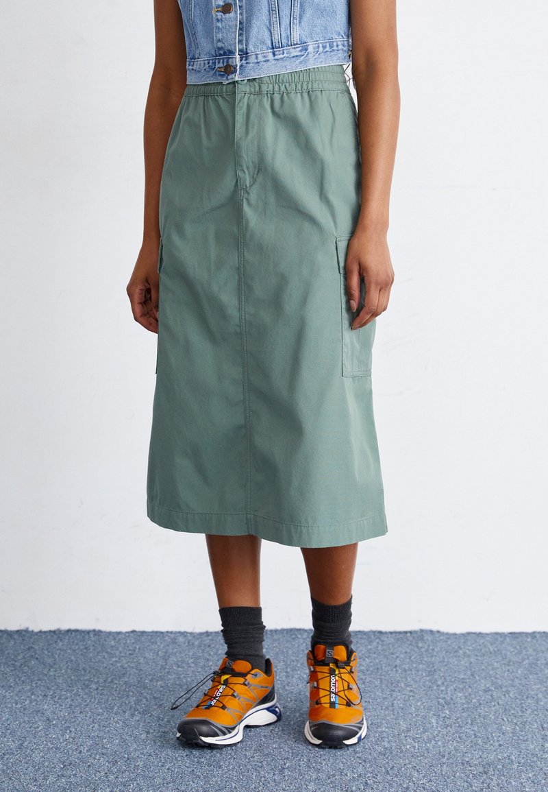 Zielona, sięgająca do połowy długości spódnica z elastycznym pasem, bocznymi kieszeniami i gładką teksturą, zestawiona z pomarańczowo-czarnymi butami sportowymi.