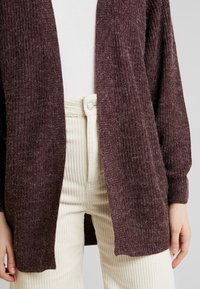 Henkilö, jolla on päällään marooninpunainen avoin neulekarvotakki valkoisen yläosan päällä, yhdistettynä korkeavyötäröisiin kermaisiin samettihousuihin.