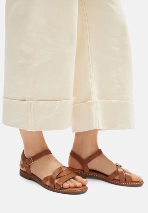 Hnědé kožené sandály s propletenými pásky a přezkou na kotníku, doplněné krémovými širokými kalhotami s roztřepenými lemy.