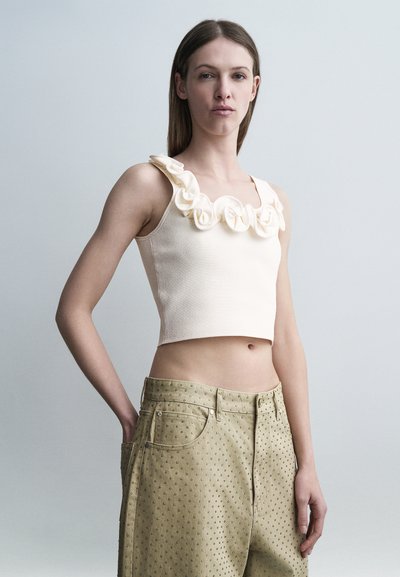 Νεαρή γυναίκα που φοράει αμάνικο μπεζ crop top με υφασμάτινα τριαντάφυλλα και μπεζ ψηλόμεσα παντελόνια με υφή πουά, στέκεται μπροστά σε γκρι φόντο.