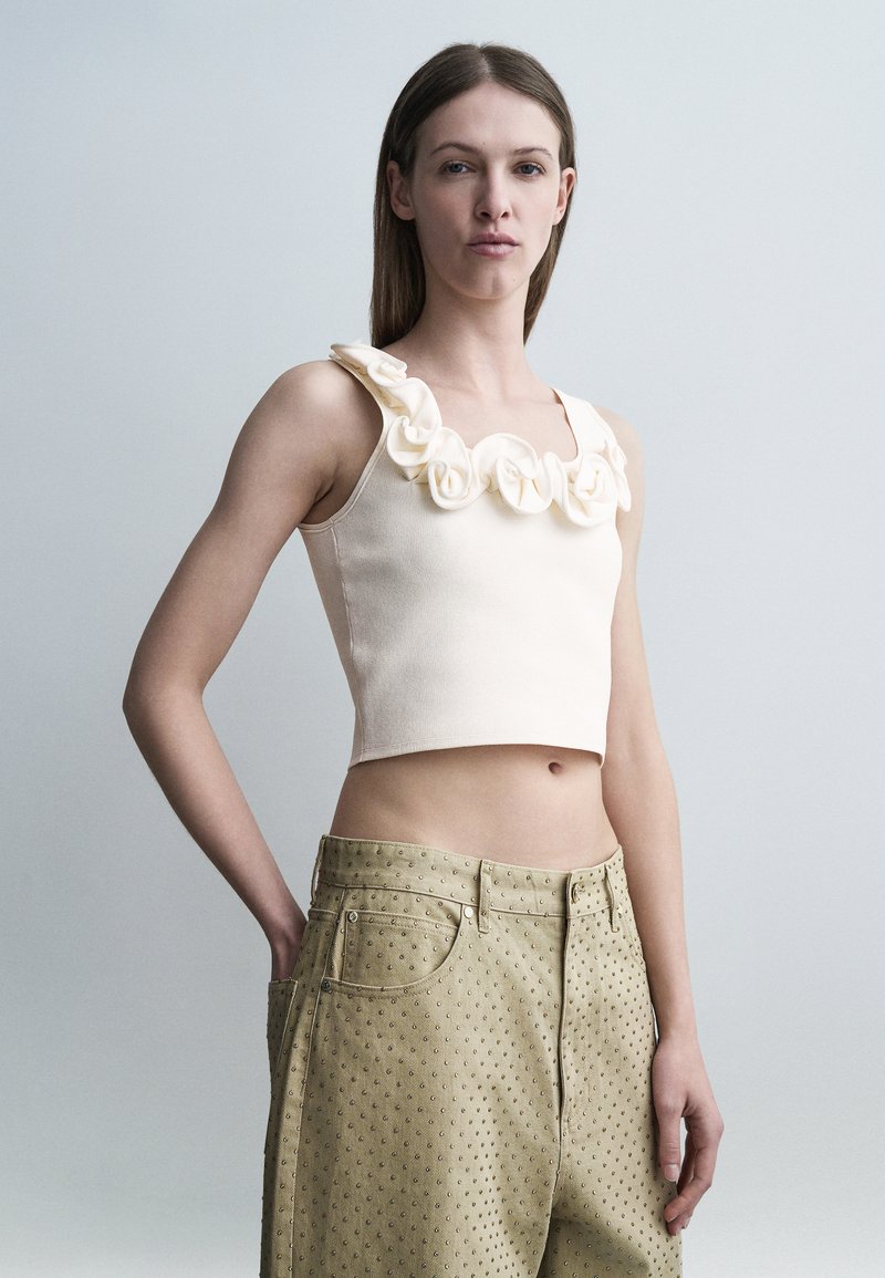 Νεαρή γυναίκα που φοράει αμάνικο μπεζ crop top με υφασμάτινα τριαντάφυλλα και μπεζ ψηλόμεσα παντελόνια με υφή πουά, στέκεται μπροστά σε γκρι φόντο.