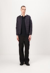 Doudoune violette foncée avec texture matelassée et accents noirs, associée à un polo noir, un pantalon cargo noir ample et des chaussures noires.