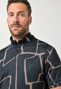 Mann mit kurzen Haaren und Bart, der ein schwarz-beiges, gemustertes Sportshirt mit Reißverschluss trägt und frontal vor einem einfarbigen Hintergrund steht.