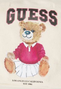 Graphique présentant un ours portant un pull rouge et une jupe blanche. L'arrière-plan est beige avec le logo "GUESS" en noir et rose.
