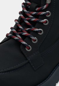 Bota negra con tobillo acolchado, que presenta cordones con un patrón rojo y azul marino, ojales plateados y un detalle de costura en contraste a lo largo de la suela.