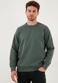 Zelený mikin s okrúhlym výstrihom, raglánovými rukávmi, ribovými manžetami a malým logom na hrudi. Mäkka textúra, voľný strih.