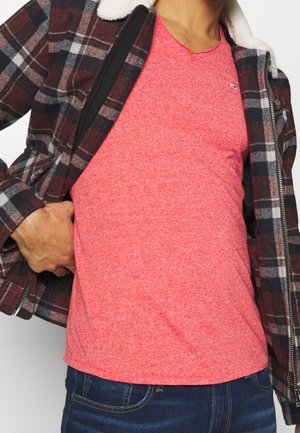 T-shirt rouge chiné à col en V sous une veste en flanelle à carreaux avec un col en peau retournée, associée à un jean bleu foncé.