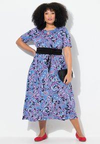 Vestido azul de paisley con mangas cortas, cintura ajustada y cinturón negro; combinado con bailarinas rojas y un bolso negro. Tela suave con patrones vibrantes.
