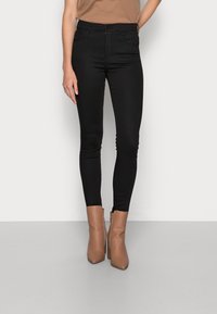 Kvinna som bär svarta högmidjade slim fit-jeans och tanfärgade ankelstövlar, står mot en enkel ljus bakgrund.