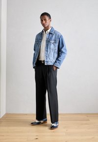 Giubbotto di jeans, blu chiaro, chiusura con bottoni frontale, due tasche sul petto, camicia bianca sottostante, pantaloni neri, scarpe blu e bianche con accenti decorativi.