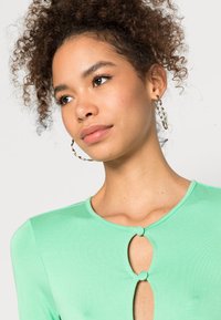 Top verde menta com decote redondo e detalhe em forma de chave segurado por um botão. Textura suave, mangas longas e design justo.
