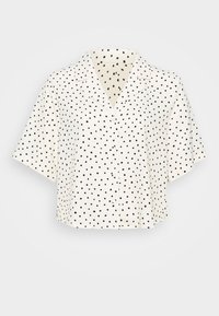 Camisa de manga corta con cuello en tejido crema con lunares negros; presenta un cierre de botones en el frente y un diseño recortado. Textura suave.