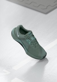Grüner Sportschuh mit einem Mesh-Obermaterial, strukturiertem Design, flachen Schnürsenkeln und einer schwarzen Laufsohle. Merkmale ein weißes Logo und kontrastierende Akzente.