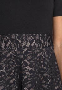 Černé šaty s přiléhavým vrškem z hladké látky a rozšířenou sukní z propracované černé krajky s květinovým vzorem.