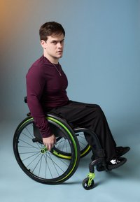 Ένα άτομο σε αναπηρικό καροτσάκι με πράσινες λεπτομέρειες, φορώντας ένα εφαρμοστό μπορντό πουκάμισο με μακριά μανίκια και μαύρο παντελόνι, σε φόντο με γραadiεντα απόχρωσης.