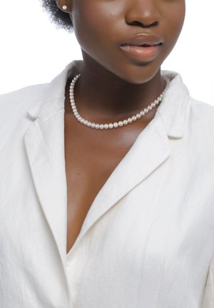 Frau trägt ein weißes Leinenblazer, Perlenkette und passende Perlensteckohrringe, mit natürlicher dunkler Haut und kurzem lockigem Haar.