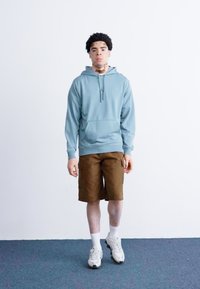 Sudadera con capucha de color azul claro con un bolsillo frontal, combinada con pantalones cortos de cargo marrones. El modelo lleva calcetines blancos y zapatillas deportivas.