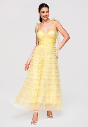 Kvinde iført en gul lagdelt tylkjole med bundne skulderstropper, smilende og poserende med den ene hånd på hoften og hvide sandaletter med hæl.