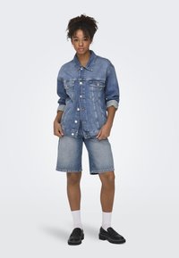 Chaqueta de mezclilla azul claro con cierre de botones y bolsillos, combinada con pantalones cortos de mezclilla a juego. El modelo lleva zapatos negros y calcetines blancos.